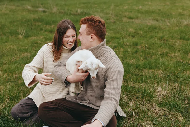 Young couple sitting on grass in casual sweaters happily holds a white baby goat enjoying a relaxed lifestyle moment outdoors in nature during autumn.