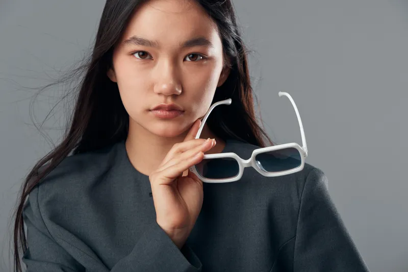 Young woman with long dark hair holding white sunglasses with serious face, wearing dark outfit against neutral background, fashion portrait, confident female model with natural look.