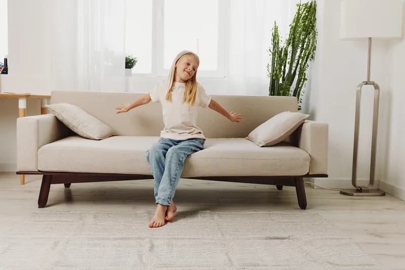 Happy young girl sitting barefoot on beige sofa in bright living room with plants, stretching arms and smiling. Concept of childhood joy, comfort, home lifestyle, and relaxation indoors.