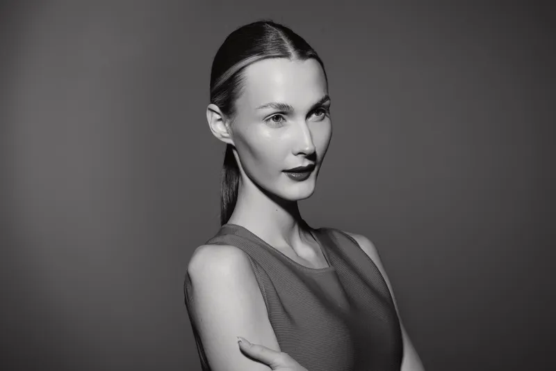 Portrait of a slender Caucasian woman with light skin and dark lipstick posing confidently against a dark background in studio lighting.