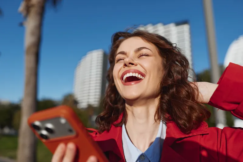 A smiling woman outdoors in a red jacket checks her phone, bright sky behind her, conveying joy, spontaneity, and casual urban lifestyle.