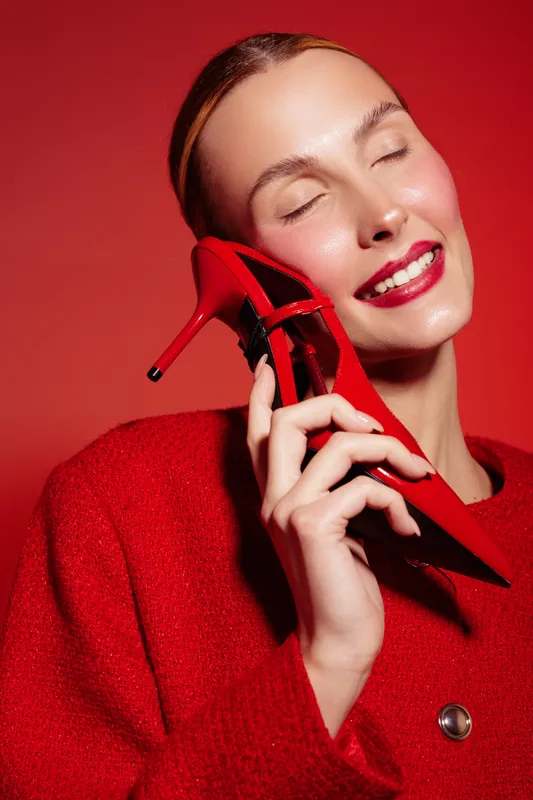 Smiling woman in red coat holds red high heel shoe near face with closed eyes. Bright studio lighting highlights glossy lips and smooth skin on red background. Concept of fashion and beauty.