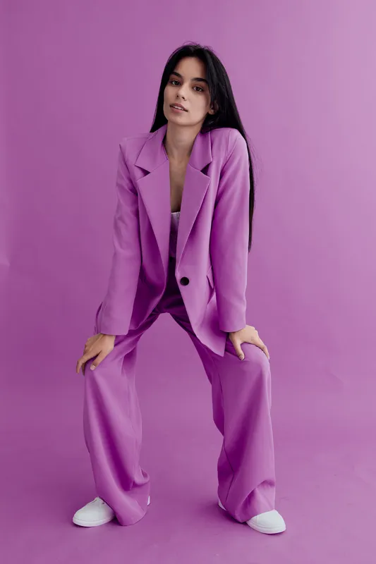 Young woman in a lavender pantsuit posing confidently against a purple backdrop, showcasing modern fashion and casual white sneakers in a studio setting.