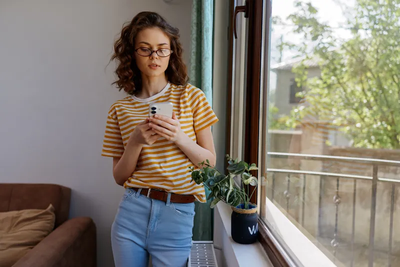 Young woman with curly hair wearing a striped tee and jeans stands by a bright window, scrolling on a smartphone in a cozy home setting, everyday moment, calm vibe, soft light