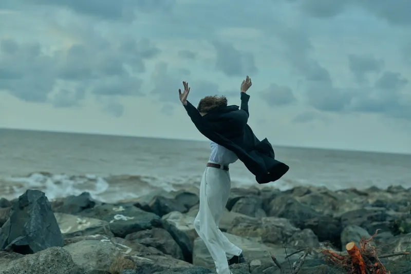A figure on a rocky coastline jumps with a flying jacket, wind swept and dramatic, capturing outdoor freedom and bold motion against a sea horizon
