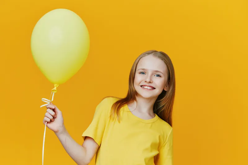 Cheerful girl in a yellow t shirt holding a yellow balloon against a bright yellow background, embodying happiness and playful joy