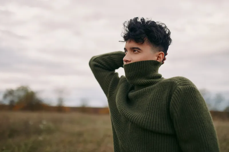 Portrait of a young man in a green turtleneck sweater standing in an open field, capturing authentic emotion and genuine presence with natural light and unposed, honest expression.