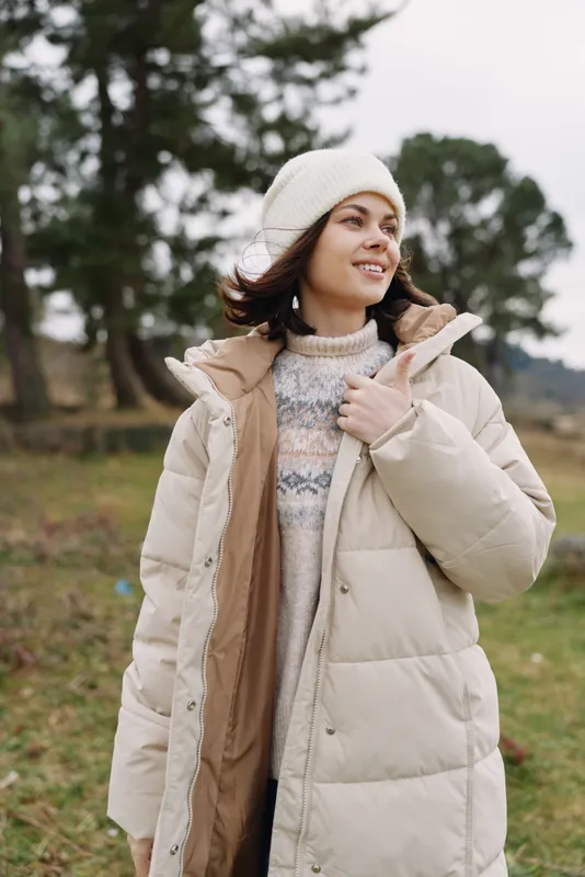 Woman wearing a puffy beige coat and knit sweater in a chilly outdoor setting, smiling softly as she adjusts her scarf and beanie.