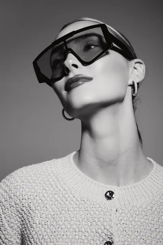 Portrait of a fair skinned Caucasian woman wearing oversized sunglasses and hoop earrings. She poses confidently in a knitted sweater against a plain background. Concept of fashion and elegance.