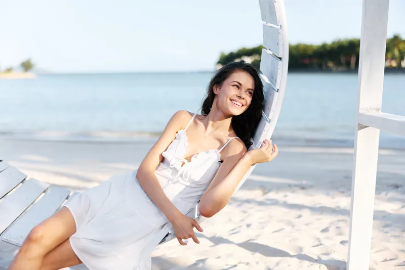 A carefree woman in a white sundress relaxes on a sunny beach, leaning against a wooden swing frame as calm waves kiss the shore.