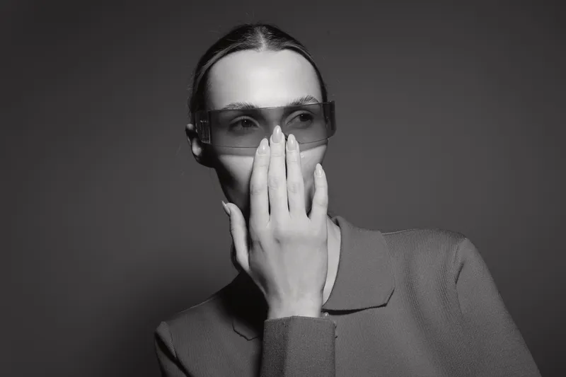 Portrait of a young Caucasian woman with pale skin wearing sunglasses and a textured jacket covering her mouth with her hand. Monochrome studio fashion concept.