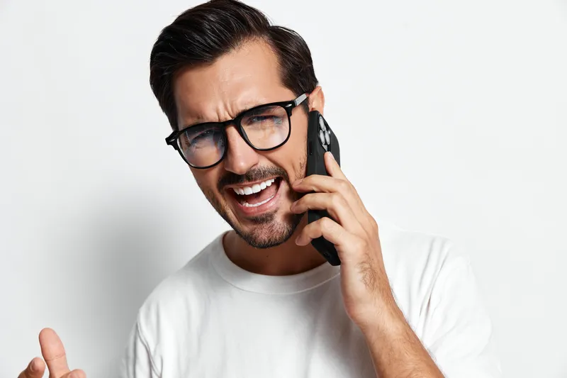 Happy smiling man talking on the phone, wearing glasses and a white T-shirt, showing excitement and engagement, isolated on plain solid light background.