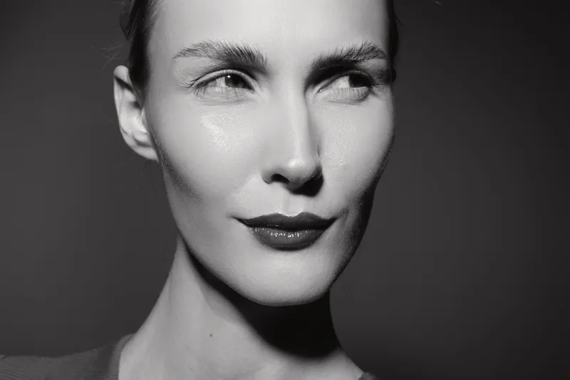 Close-up portrait of a white woman with pale skin and slicked-back hair looking thoughtfully to the side. Black and white studio lighting highlights facial features and expression.