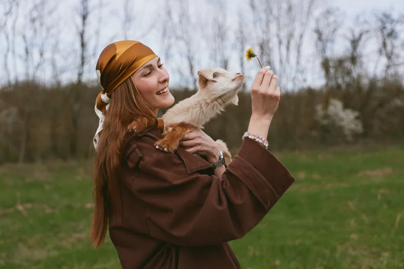 Cheerful woman outdoors holding a playful goat kid, offering a yellow flower, warm tones, casual jacket, natural light, relaxed pose, countryside setting, lifestyle moment with animals