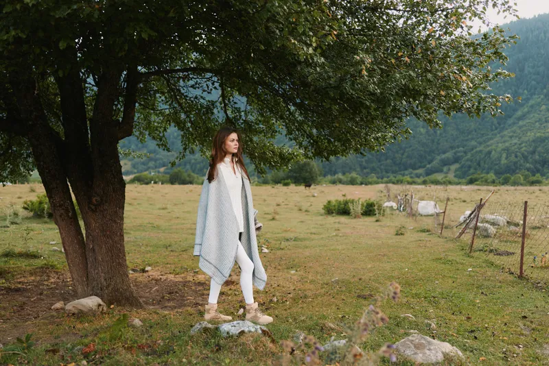 Young woman walks through a sunlit countryside field beside a large tree, wearing a light gray coat and white outfit, a calm contemplative mood