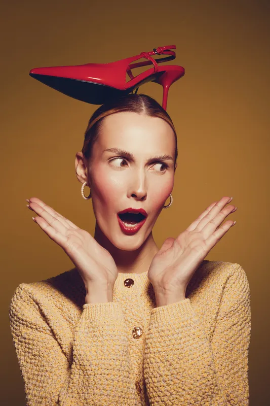Surprised woman with red high heel shoe balanced on her head, wearing yellow textured jacket and gold hoop earrings. Concept for fashion, style, and playful attitude in studio.