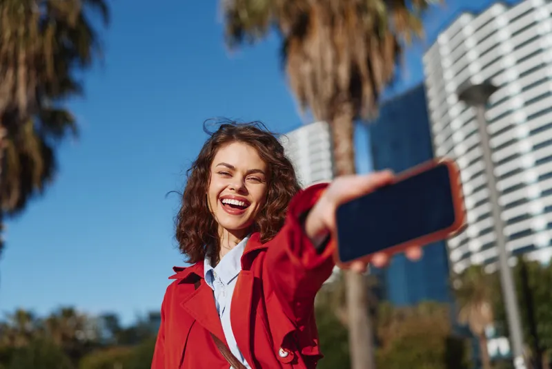 A smiling woman takes a selfie in a sunny urban setting with palm trees, wearing a red jacket. A vibrant lifestyle moment for outdoor, travel, and selfie photography.