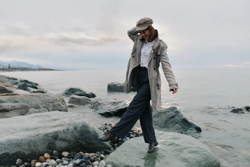 A stylish woman in a long coat and hat walks on rocky shore near calm sea under cloudy sky. The scene conveys fashion, nature, and autumn mood with casual elegance and outdoor lifestyle.