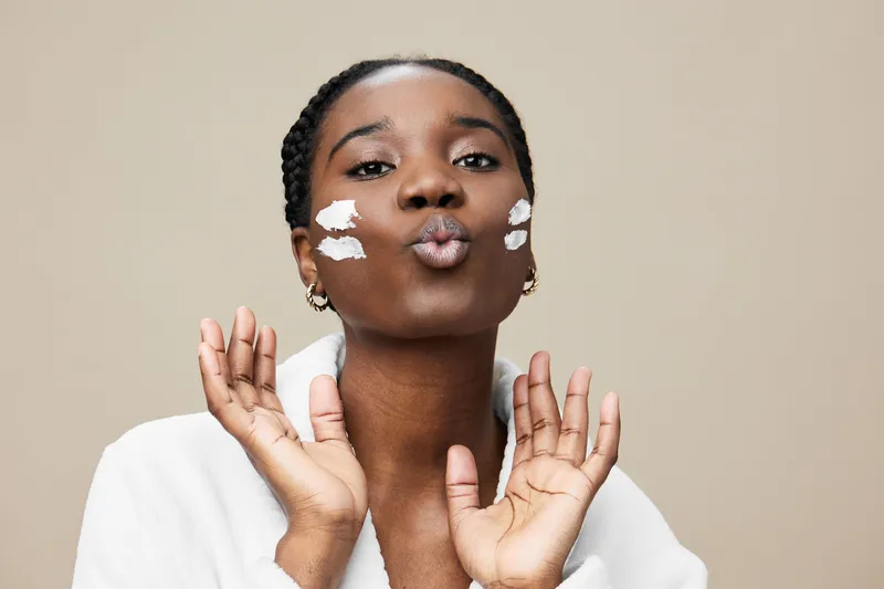 Young black woman applying facial cream with a playful expression, showcasing a natural skincare routine against a soft beige background