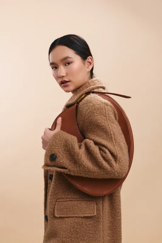Asian woman wearing a textured coat carries a round leather backpack, posing against a soft studio backdrop. The image conveys style, confidence, and modern fashion with a warm, minimal palette.