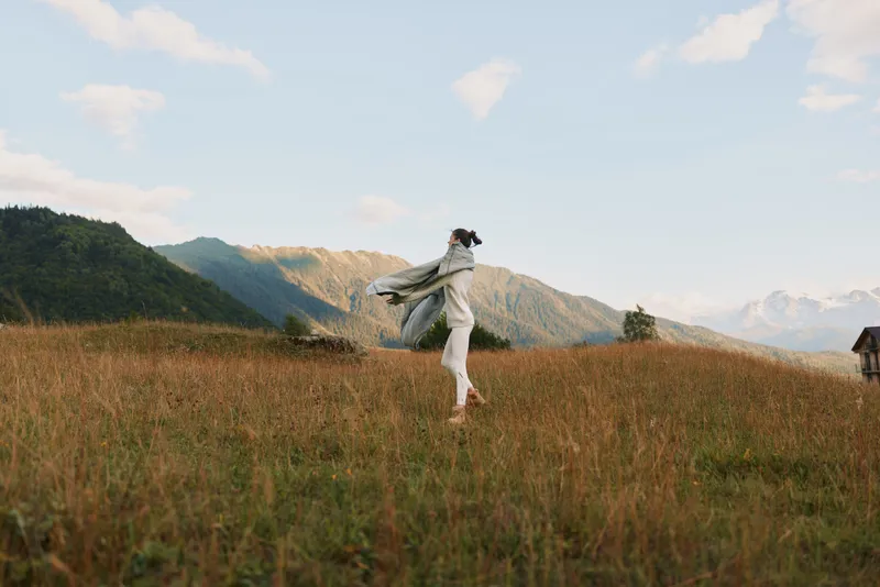 Young woman in casual clothes enjoys freedom and fresh air running through grassy field with mountains in background under blue sky with clouds.