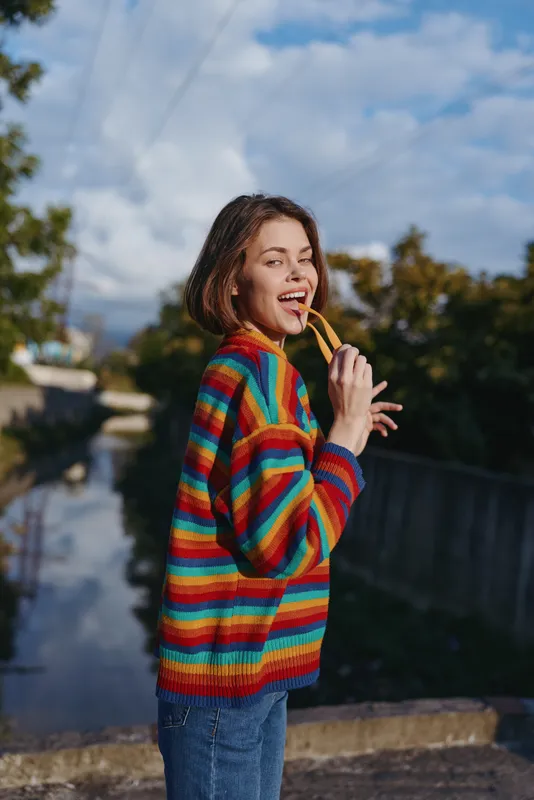 Young woman in her early twenties wearing a colorful striped sweater and casual outfit, smiling and playful while holding sunglasses by a canal outdoors for lifestyle travel portrait.