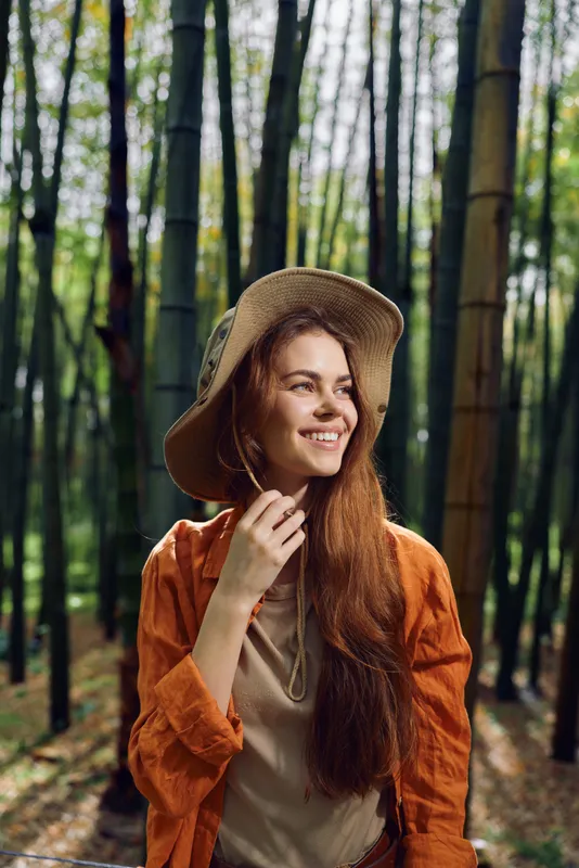 Woman smile hat bamboo forest nature portrait outdoor, young redhead woman in orange jacket wearing wide brim hat, smiling and enjoying sunlit bamboo grove during a relaxed nature walk.