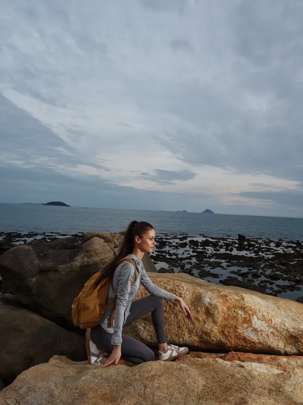 Young woman enjoying outdoor adventure on rocky beach, wearing sportswear and backpack, against serene coastal background with cloudy sky, emphasizing nature and fitness