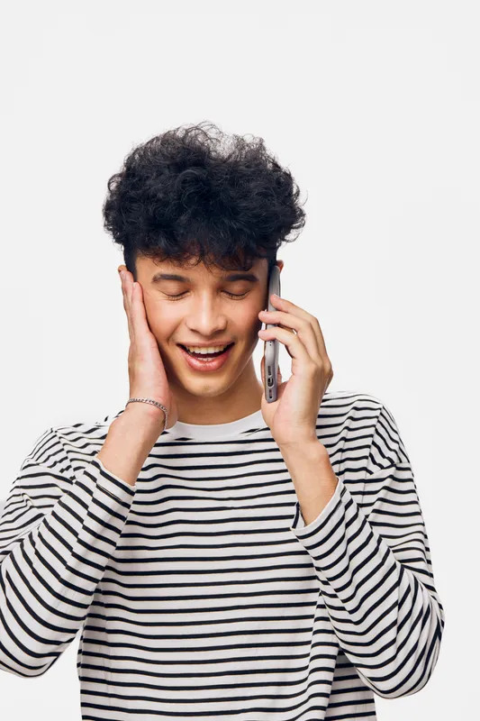 smiling young man in striped shirt talking on phone, expressing joy with hand on face, isolated white background, casual communication moment