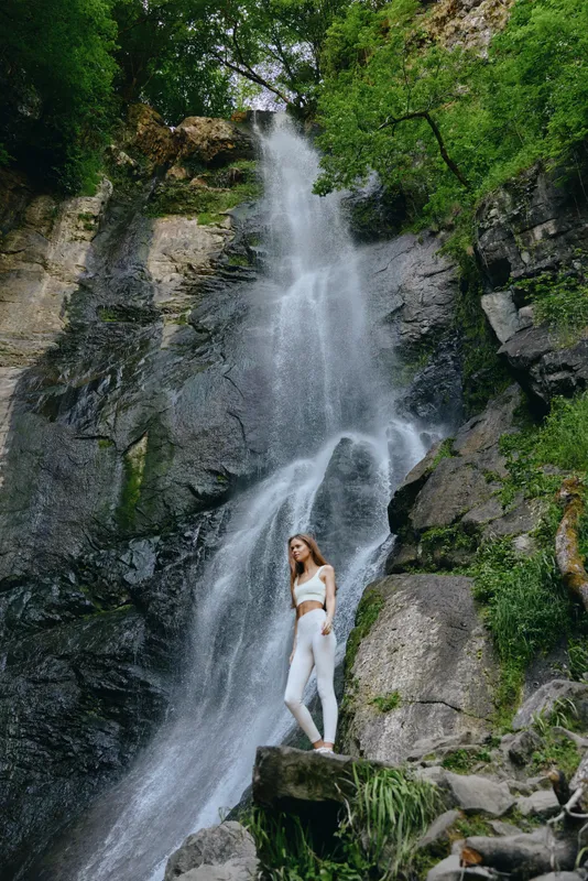 woman standing near waterfall in white activewear, surrounded by lush green forest and rocky cliffs, showing outdoor adventure and natural beauty.