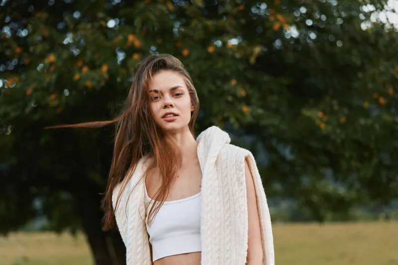 Young woman outdoors in a white crop top with a knit cardigan draped over her shoulders. Bright natural light, park surroundings, casual lifestyle moment.
