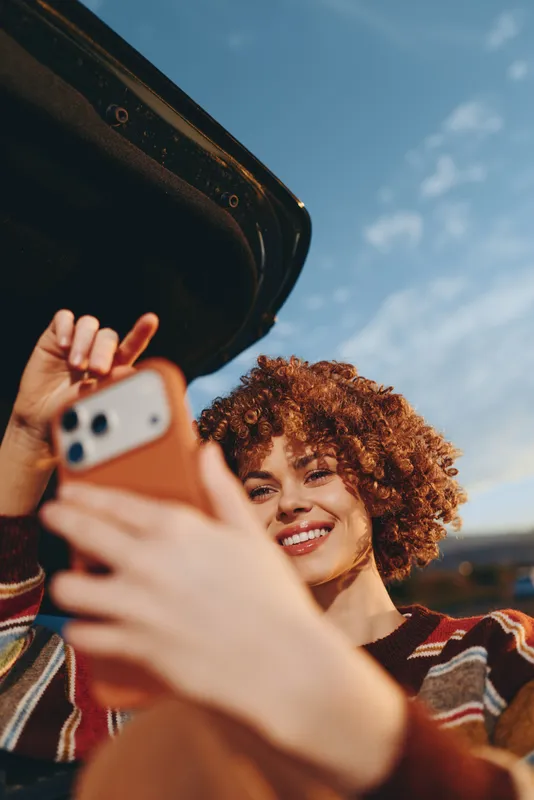 Smiling woman with curly hair takes a selfie with smartphone outdoors at golden hour, warm lifestyle moment, hands holding phones in foreground, sunny sky background, casual street fun