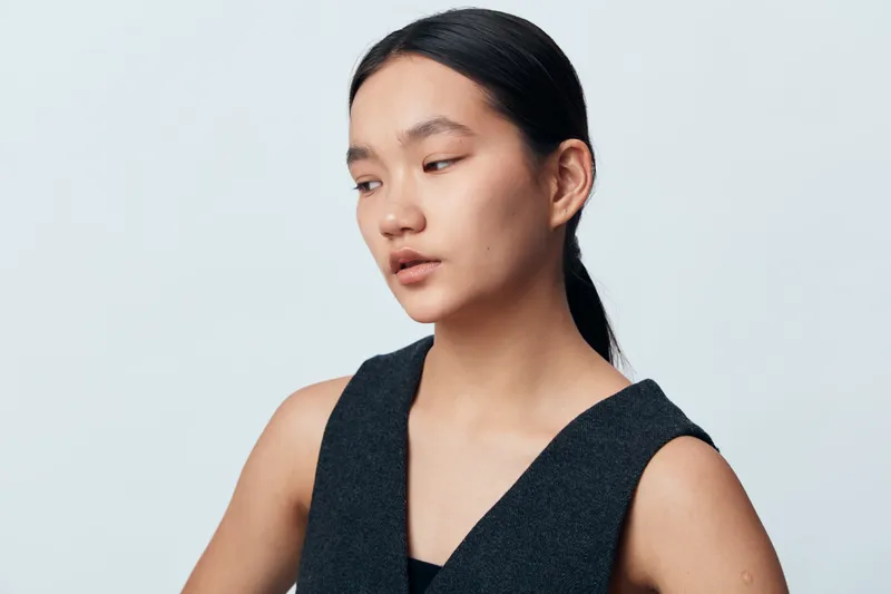 Portrait of young woman with black hair and natural makeup looking aside on white background. Asian female model in casual dark sleeveless top with smooth skin and neutral expression.