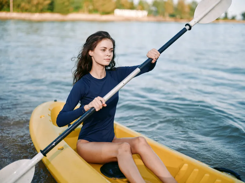 Focused paddler sits in a yellow kayak on calm water, gripping the paddle with determination, wind tousling hair, and sunlight highlighting a navy top for a vivid sport moment.