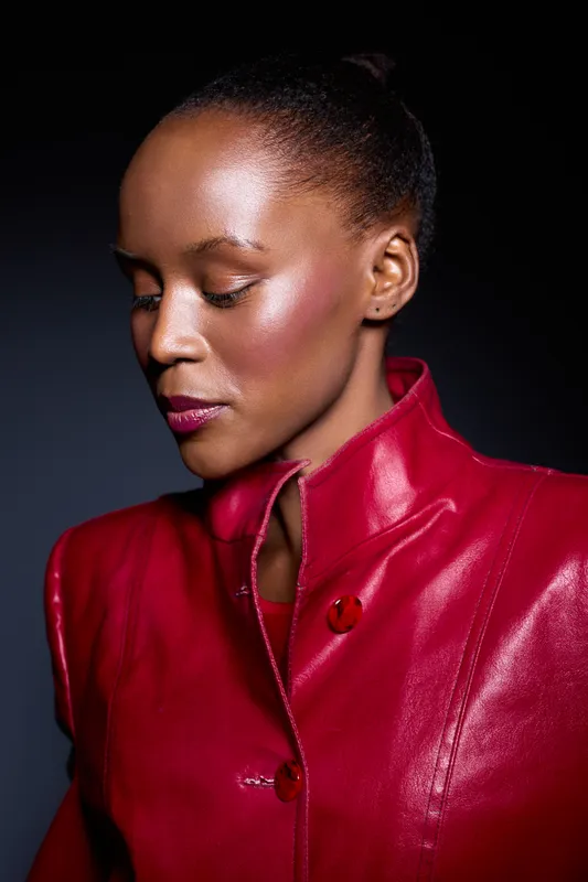 Portrait of a thoughtful African woman with dark skin in a shiny red leather jacket against dark background. Concept for fashion, beauty, elegance, and confidence in studio lighting.