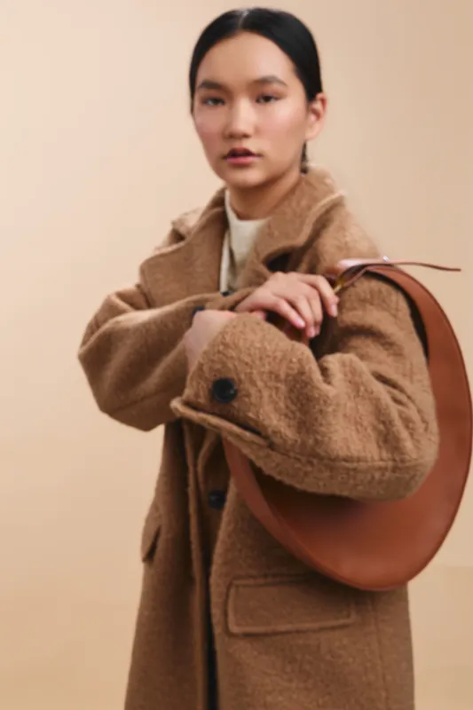 Asian woman in a warm coat stands confidently with a bag, expressing poise and style in a minimal studio setting, inviting curiosity and elegance.