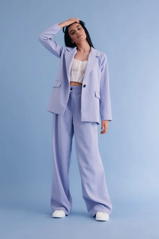 young woman in lilac suit posing confidently with hand on forehead against pastel blue background, wearing white lace top and sneakers, stylish outfit