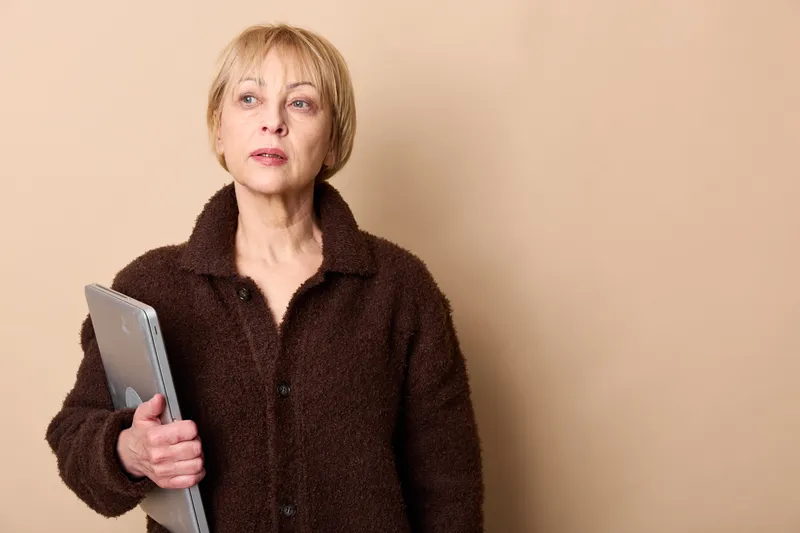 Elderly woman in dark coat holding tablet, studio portrait on beige background, calm expression, looking at camera, using digital technology for communication, today