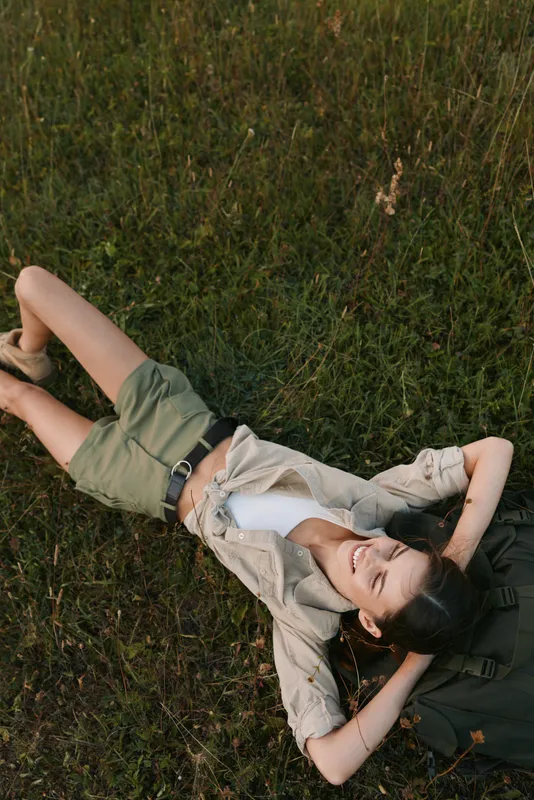 A relaxed woman lies on grass in casual summer clothes, smiling with arms overhead. An outdoor lifestyle scene conveying calm, leisure, and carefree moments.