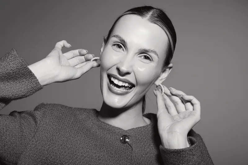Happy Caucasian woman with fair skin and slicked back hair smiling broadly while holding earrings near her ears on grey background. Concept of joy and fashion accessory.