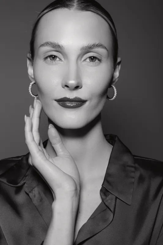 Close-up portrait of a fair skinned Caucasian woman with slicked hair and hoop earrings, wearing a satin blouse and touching face.