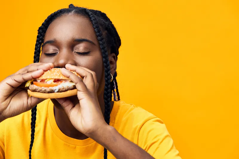 Young African woman enjoying a delicious burger, eyes closed, wearing a bright yellow shirt, against a vibrant yellow background, perfect for food ads