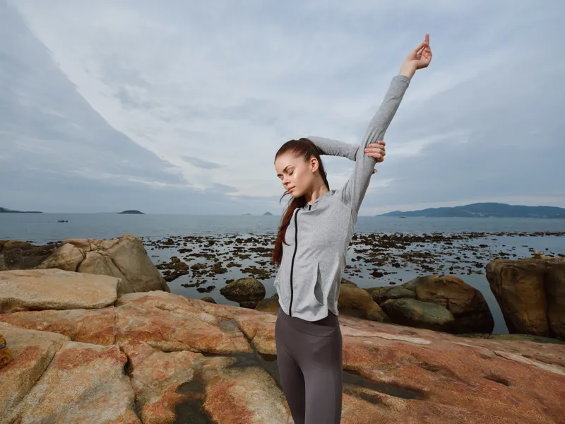 Young athletic woman stretching on rocky beach at dawn, wearing sportswear, expressing determination and focus, with a serene ocean view in the background