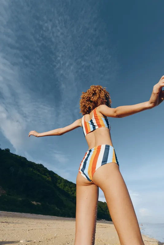 Woman in swimsuit with curly hair on beach, arms extended toward ocean under clear blue sky, carefree summer vacation concept, rear view, sunlight