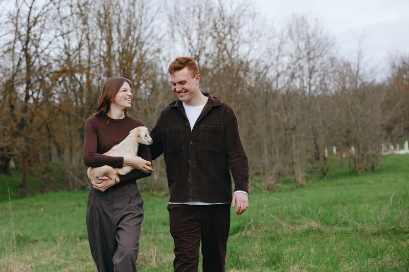 Smiling couple walking with a goat kid in a grassy park, enjoying a relaxed countryside moment. Outdoor lifestyle scene showing companionship, nature, and gentle animal interaction together.