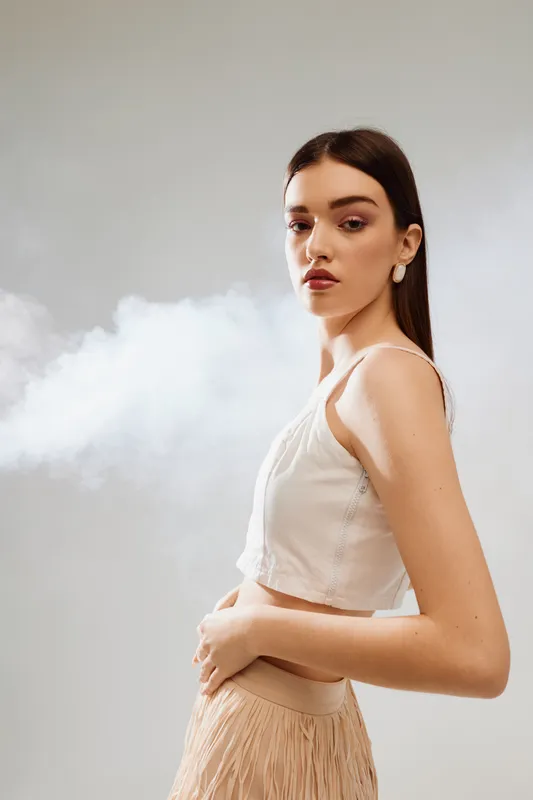 Young woman in white top and beige skirt poses with confident expression against light background with soft smoke. Concept of beauty fashion and elegance in studio.
