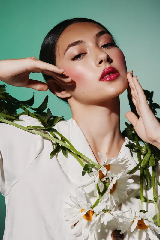 Elegant young woman with flawless skin poses with flower stems in hands, wearing bright makeup and white attire against green background. Artistic beauty creative fashion concept.