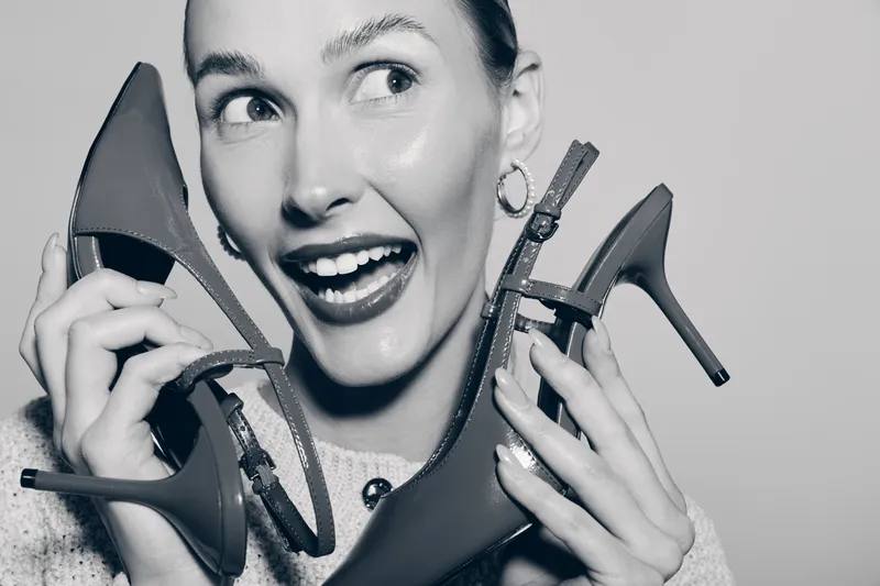 Happy Caucasian woman with light skin holds high heel shoes near face, smiling excitedly. Concept for fashion, footwear, shopping, and lifestyle in studio lighting.