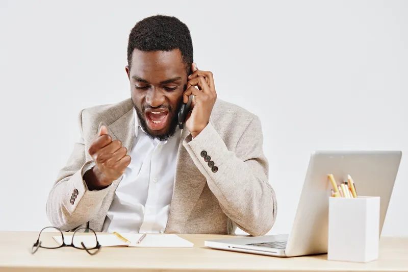 Professional young African American man in formal beige blazer and white shirt talking on mobile phone while working at office desk with laptop and glasses, excited expression, indoors. Business commu