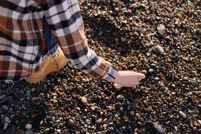Pebbles on a rocky beach with a person kneeling and picking stones, casual plaid sleeve visible, outdoor texture and exploration of the shoreline.